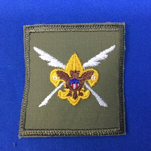 Op de afbeelding: Een groene stoffen patch met een gele geborduurde fleur-de-lis, een bruine adelaar en een wit schild met rode en blauwe accenten. Twee witte gekruiste pijlen bevinden zich achter de fleur-de-lis.