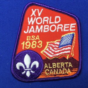 May include: A red, white, and blue embroidered patch with the text "XV WORLD JAMBOREE BSA 1983 ALBERTA CANADA". The patch features a white fleur-de-lis on a purple circle and a stylized American flag.