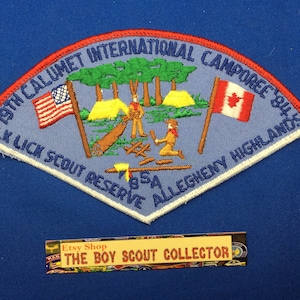 Może przedstawiać: Naszywka harcerska w kolorze niebieskim i białym z czerwonym i białym napisem. Naszywka przedstawia scenę z dwoma harcerzami obozującymi w lesie z namiotem, ogniskiem i kłodą. Tekst na naszywce brzmi "19th Calumet International Camporee 84 Elk Lick Scout Reserve BSA Allegheny Highlands".