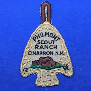 Op de afbeelding: Een beige geborduurde patch in de vorm van een pijlpunt met de tekst "Philmont Scout Ranch Cimarron N.M." in zwarte letters. De patch toont een bruin bergketen met groen gras ervoor.