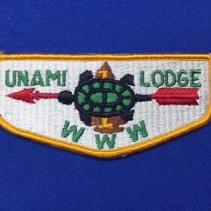 Puede incluir: Un parche blanco con borde amarillo, que presenta una tortuga verde con caparazón negro y una flecha roja apuntando hacia la izquierda. El texto "UNAMI LODGE" está escrito en letras verdes sobre la tortuga, y "WWW" está escrito en letras verdes debajo de la tortuga.