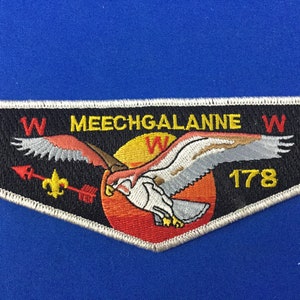 Puede incluir: Un parche bordado blanco, negro y amarillo con un águila volando, un sol y el texto "W MEECHGALANNE W 178".