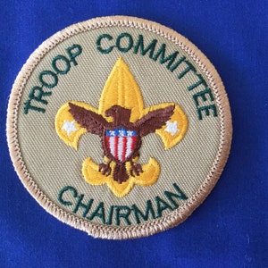 May include: A tan and green embroidered patch with the text "Troop Committee Chairman" and a gold and brown scout emblem.