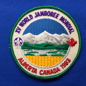 Op de afbeelding: Ronde geborduurde patch met een berg- en meerlandschap. De patch bevat de tekst "IV WORLD JAMBOREE MONDIAL", "ALBERTA CANADA 1983" en een lelie-symbool. Het ontwerp toont een esdoornblad en een groene rand.
