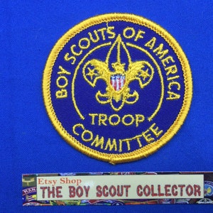 Op de afbeelding: Een paarse en gouden Boy Scouts of America Troop Committee-badge. De badge heeft het Boy Scouts of America-logo met de woorden "Boy Scouts of America" en "Troop Committee" rond het logo.