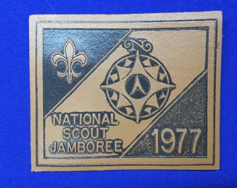 Puede incluir: Un parche de cuero marrón con letras negras y un diseño de brújula. El texto dice "National Scout Jamboree 1977".