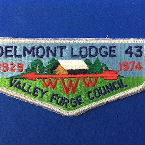 Puede incluir: Un parche bordado azul y verde con el texto "Delmont Lodge 43" y "Valley Forge Council". El parche también presenta una flecha roja con una pequeña casa blanca y negra en el fondo.