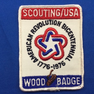 Puede incluir: Un parche blanco de Scouting/USA con un diseño de estrella rojo, blanco y azul. El texto "American Revolution Bicentennial 1776-1976" está alrededor de la estrella. El texto "Wood Badge" está en la parte inferior del parche.