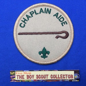 May include: A beige round patch with green embroidered text that reads "Chaplain Aide". The patch features a brown shepherd's crook and a green embroidered scout emblem.