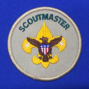 May include: A tan Scoutmaster patch with a green embroidered border. The patch features a yellow fleur-de-lis with a brown eagle and a red, white and blue shield. The words "Scoutmaster" are embroidered in green around the edge of the patch.