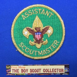 May include: A circular embroidered patch with a green background and a gold border. It features a gold fleur-de-lis emblem with a brown eagle and a red, white, and blue shield. The text reads ASSISTANT SCOUTMASTER in gold thread.