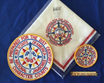 Pode incluir: Um lenço de escoteiro branco com guarnições vermelho, branco e azul. O lenço tem o texto "Be Prepared for the 21st Century Boy Scouts of America National 5 Jamboree Character Counts" impresso nele. Existem dois patches redondos bordados com o mesmo design, um maior e outro menor. Os patches são amarelos, vermelhos, azuis e brancos com uma bússola e o texto "1937 1997".
