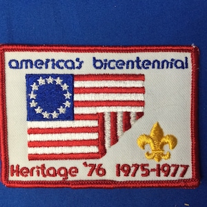 Puede incluir: Un parche bordado blanco con costuras rojas y azules. El parche presenta una bandera estadounidense estilizada con 13 estrellas, una flor de lis y el texto "america's bicentennial Heritage '76 1975-1977".