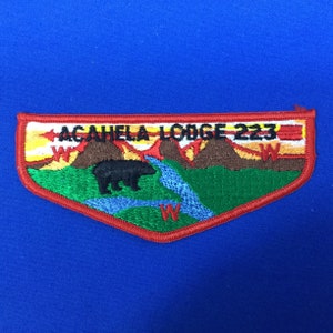 Puede incluir: Un parche bordado en rojo, blanco y verde con el texto "AGAHELA LODGE 223". El parche presenta un oso negro, un río azul y montañas marrones.