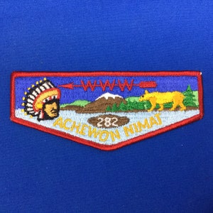 Puede incluir: Un parche de Boy Scout azul y rojo con un jefe nativo americano, una montaña, un lobo y el texto "Achewon Nimat 282".