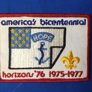 Puede incluir: Un parche blanco bordado con ribete rojo. El parche presenta un ancla azul, una pancarta azul con la palabra "HOPE" en blanco, y una bandera azul y dorada con 13 estrellas. El texto "america's bicentennial" está por encima de la bandera y el texto "horizons '76 1975-1977" está por debajo de la bandera. Una flor de lis dorada está a la derecha de la bandera.