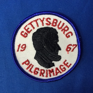 May include: A white circular patch with red and blue trim. The patch features a black silhouette of a man's head and the text "Gettysburg 1967 Pilgrimage".