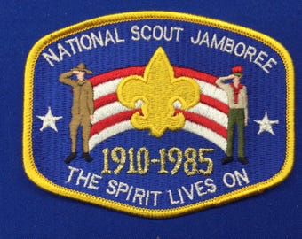 Pode incluir: Um remendo bordado azul e dourado com o texto "National Scout Jamboree 1910-1985 The Spirit Lives On". O remendo apresenta uma flor-de-lis amarela, duas figuras em uniforme a saudar e uma faixa listrada vermelha, branca e azul.