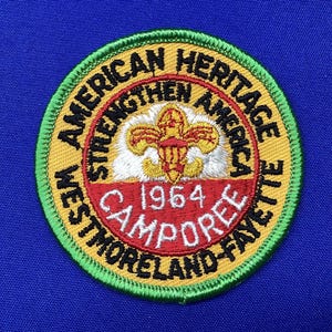Może przedstawiać: Haftowana naszywka z napisem "American Heritage" i "Westmoreland-Fayette Camporee 1964". Naszywka ma złotą, czerwoną i zieloną kolorystykę ze skautowskim emblematem pośrodku. Naszywka jest okrągła.