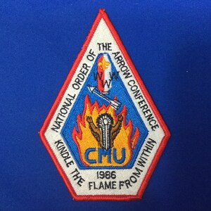 Puede incluir: Un parche blanco en forma de diamante con ribete rojo y azul. El parche presenta una figura nativa americana sosteniendo una antorcha en llamas con una flecha volando a través de las llamas. El texto dice "National Order of the Arrow Conference Kindle the Flame From Within 1986".