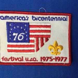 Puede incluir: Un parche blanco bordado con una bandera estadounidense roja, blanca y azul, el número "76" en azul, una flor de lis amarilla y el texto "america's bicentennial festival u.s.a. 1975-1977".