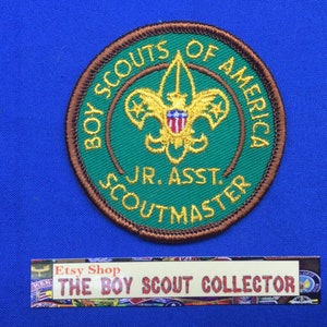 May include: A green and brown Boy Scouts of America patch with the text "Boy Scouts of America" and "Jr. Asst. Scoutmaster" around a gold embroidered scout emblem.