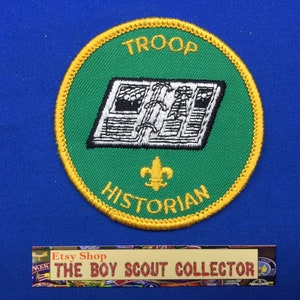 Op de afbeelding: Een groene en gouden Boy Scout Troop Historian badge met een boek en fleur-de-lis ontwerp.