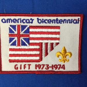 Puede incluir: Un parche blanco bordado con costuras rojas y azules. El parche presenta una bandera con 13 rayas y una Union Jack, junto con el texto "America's Bicentennial GIFT 1973-1974".
