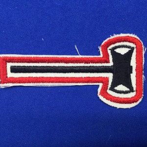 May include: A red, white, and black embroidered patch with a stylized axe design. The axe is black with a white outline and is set against a red and white striped background.