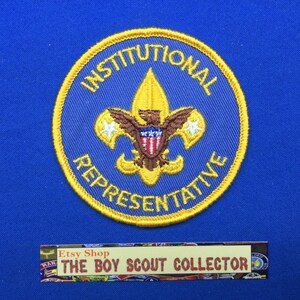 Op de afbeelding: Een blauwe en gouden geborduurde scoutingbadge met de tekst "Institutionele vertegenwoordiger" en een gouden geborduurd scoutingembleem in het midden.