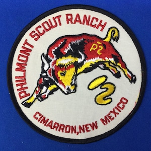 May include: A white, round embroidered patch with a black border. The patch features a red and yellow bull with a black outline, jumping over a yellow number "2". The text "Philmont Scout Ranch Cimarron, New Mexico" is written in black around the bull.