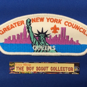 May include: Embroidered patch featuring the Statue of Liberty, a skyline, and the text "GREATER NEW YORK COUNCILS QUEENS." The patch has a white border, with a blue, purple, and orange color scheme. The patch is on a blue background.