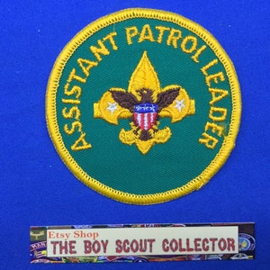 Op de afbeelding: Een cirkelvormige groen en goud geborduurde patch met de gele woorden "ASSISTANT PATROL LEADER". De patch heeft een gouden fleur-de-lis en een adelaar met een schild. De patch staat op een blauwe achtergrond.
