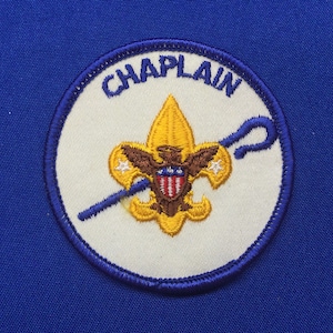 Op de afbeelding: Een ronde witte en blauwe patch met het woord "CHAPLAIN" in blauwe letters. De patch heeft een geborduurd Scout-embleem in geel met een bruine adelaar en een rood-wit-blauw schild.