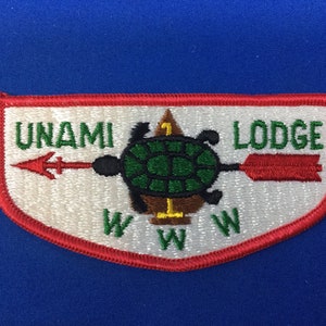 Puede incluir: Un parche blanco con borde rojo, que presenta una tortuga verde con caparazón marrón y una flecha roja apuntando hacia la derecha. El texto "UNAMI LODGE" está escrito en letras verdes en el parche.