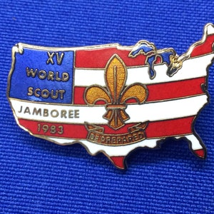 Puede incluir: Un pin de color dorado con forma de Estados Unidos con un diseño de bandera americana roja, blanca y azul. El pin presenta el texto "XV WORLD SCOUT JAMBOREE 1983" y una flor de lis dorada con el texto "BE PREPARED" debajo.