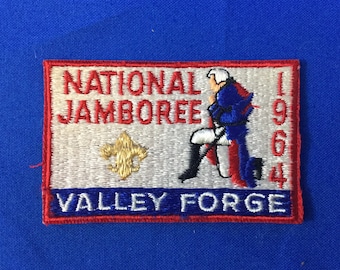 Pode incluir: Um remendo bordado branco e vermelho com o texto "NATIONAL JAMBOREE 1964 VALLEY FORGE". Uma imagem de desenho animado de um homem com um uniforme azul e vermelho está ajoelhado do lado direito do remendo. Um emblema de escoteiro dourado está do lado esquerdo do remendo.