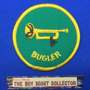 Op de afbeelding: Een ronde groene en gele Boy Scout-badge met een gouden trompet en het woord "BUGLER" in gele letters.