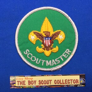 May include: A green and white embroidered Scoutmaster badge featuring a yellow fleur-de-lis and an eagle with a shield. The text "Scoutmaster" is written in a white arc around the fleur-de-lis.
