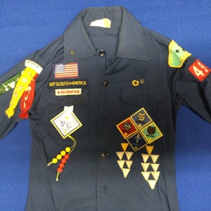 Puede incluir: Camisa de uniforme azul marino de los Boy Scouts of America con múltiples parches e insignias. Cuenta con un parche de la bandera estadounidense, un parche de "Recruiter" y varias insignias de mérito. Incluye un cordón de cuentas y un parche "41".