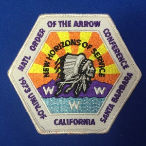Puede incluir: Un parche hexagonal blanco con el texto "NATL ORDER OF THE ARROW CONFERENCE 1973 UNIV. OF CALIFORNIA SANTA BARBARA" que rodea una imagen colorida de un tocado nativo americano con el texto "NEW HORIZONS OF SERVICE" en un círculo a su alrededor.