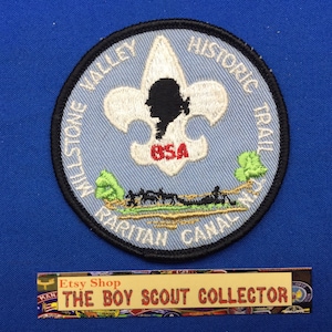 May include: A circular patch with a black border, featuring a light blue background with white text that reads "Millstone Valley Historic Trail Raritan Canal." The patch includes a silhouette and the word "USA".