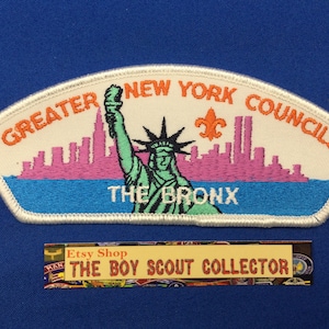 May include: A white embroidered Boy Scout patch with the text "GREATER NEW YORK COUNCILS" and "THE BRONX". The patch features a green Statue of Liberty with an orange torch, a pink cityscape, and a blue water scene.
