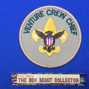 May include: A tan and green Venture Crew Chief patch with a yellow fleur-de-lis and an eagle with a shield featuring the American flag.