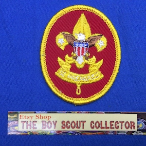 May include: A circular Boy Scout patch with a red background and a gold border. The patch features a gold fleur-de-lis, an eagle, and a banner with text. The patch is on a blue background. The text "THE BOY SCOUT COLLECTOR" is visible.