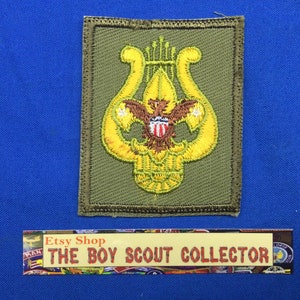 Op de afbeelding: Een groene stoffen patch met een gele geborduurde lier en een adelaar. De adelaar heeft een rood, wit en blauw schild op zijn borst. De patch is voor de Boy Scouts of America.