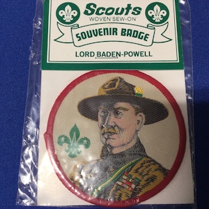 Könnte beinhalten: Ein rundes, gesticktes Scout-Souvenir-Abzeichen mit einem Porträt von Lord Baden-Powell in Uniform. Das Abzeichen hat einen roten Rand und einen weißen Hintergrund mit einem grünen Scout-Emblem.