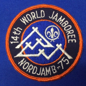May include: Embroidered patch with the text "14th WORLD JAMBOREE" in white on a navy blue background, with an orange border. The patch features a mountain design and a fleur-de-lis symbol. The text "NORDJAMB-75" is also included.