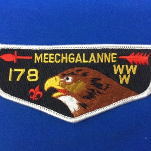 Puede incluir: Un parche bordado en blanco y negro con una flecha roja y amarilla, una lanza roja y amarilla y el texto "MEECHGALANNE 178 WWW". Una cabeza de águila marrón y blanca está en el centro del parche.
