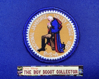 Pode incluir: Remendo bordado circular com uma figura ajoelhada em um casaco azul. O remendo tem o texto "National Jamboree Valley Forge 1950 B.S.A." em um fundo azul.